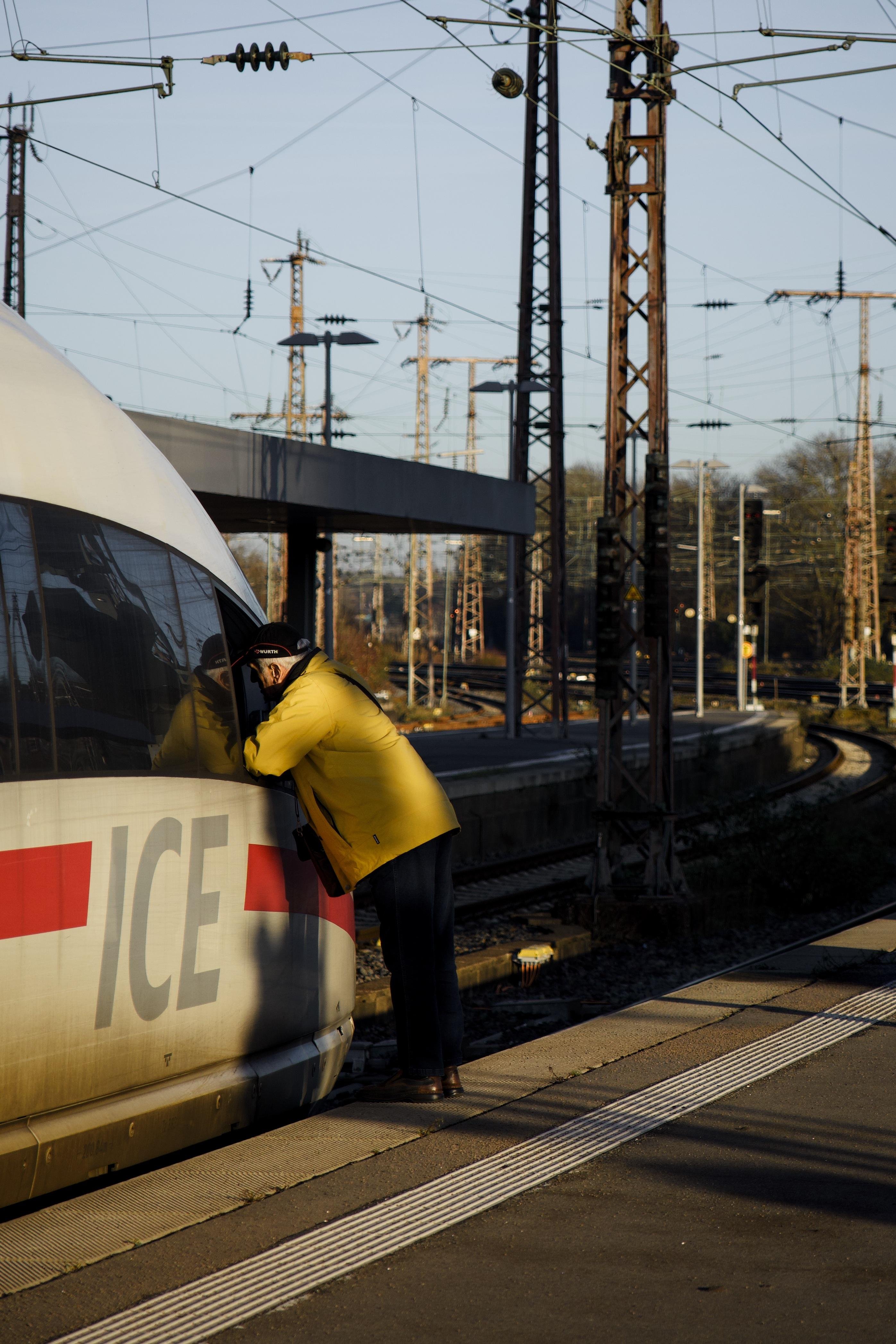 The image depicts a person in a yellow jacket leaning against the window of a train marked with "ICE" at a railway station. There are overhead wires and tracks visible in the background, indicating it's a train station setting.