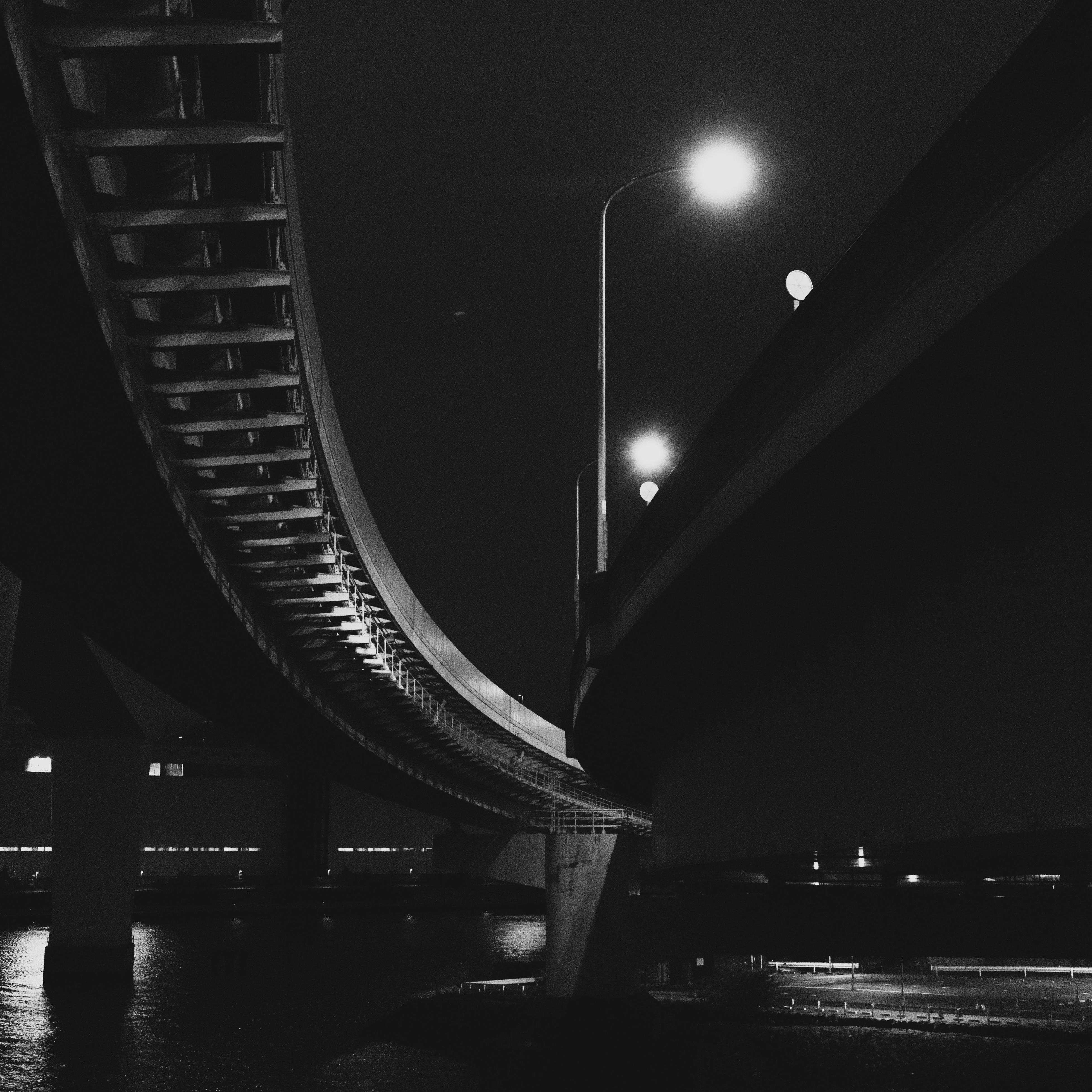 Two elevated highways curving in opposite directions at night. Streetlights illuminate the scene, casting light on the road surfaces. The structures are supported by columns, and a dark water body is visible below. The image is in black and white, emphasizing contrast and shadows.