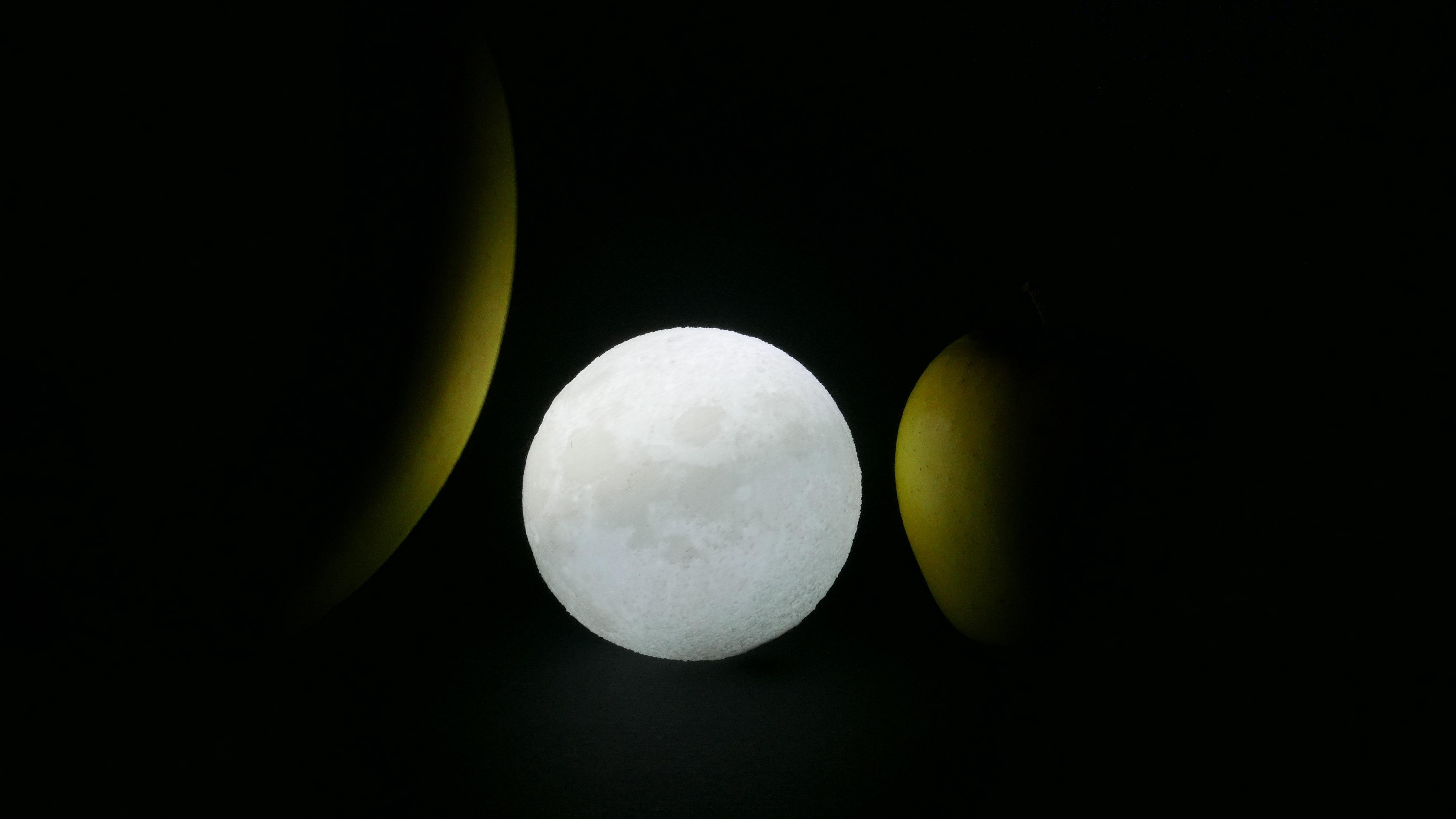 The image shows a round, glowing object resembling the moon placed between two bananas. The setup creates an interesting visual contrast with the bright moon-like object and the darker bananas on either side.