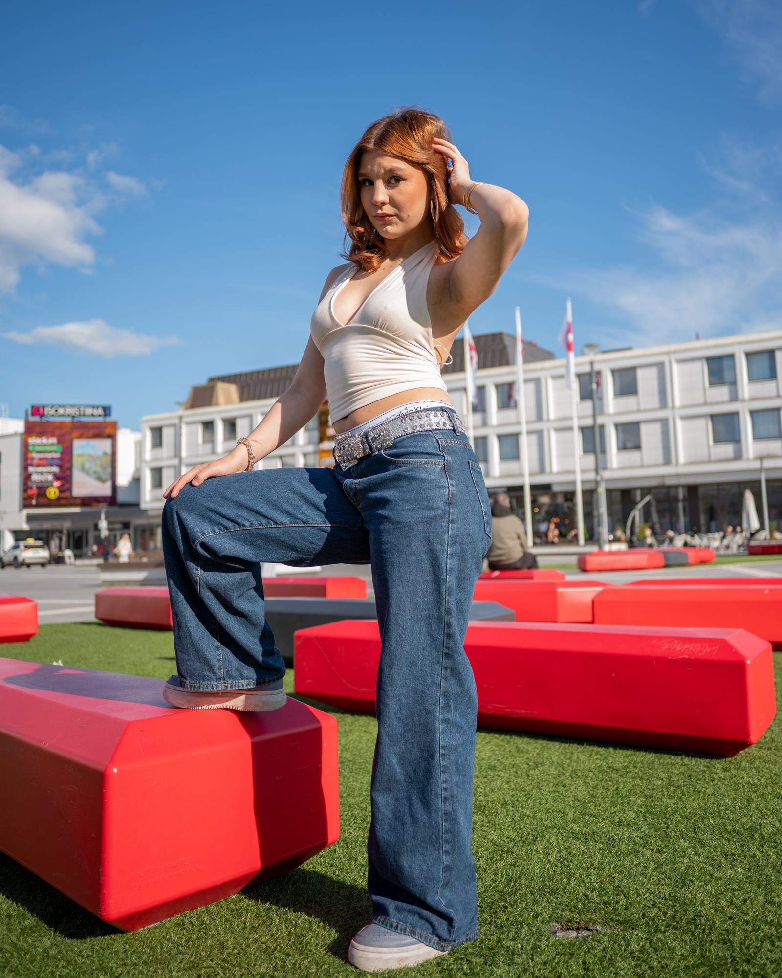 Woman with red hair wearing a white sleeveless top and blue jeans poses with one foot on a large red rectangular block. Background features a modern building and clear blue sky. Bright sunlight casts shadows on the artificial grass surface.
