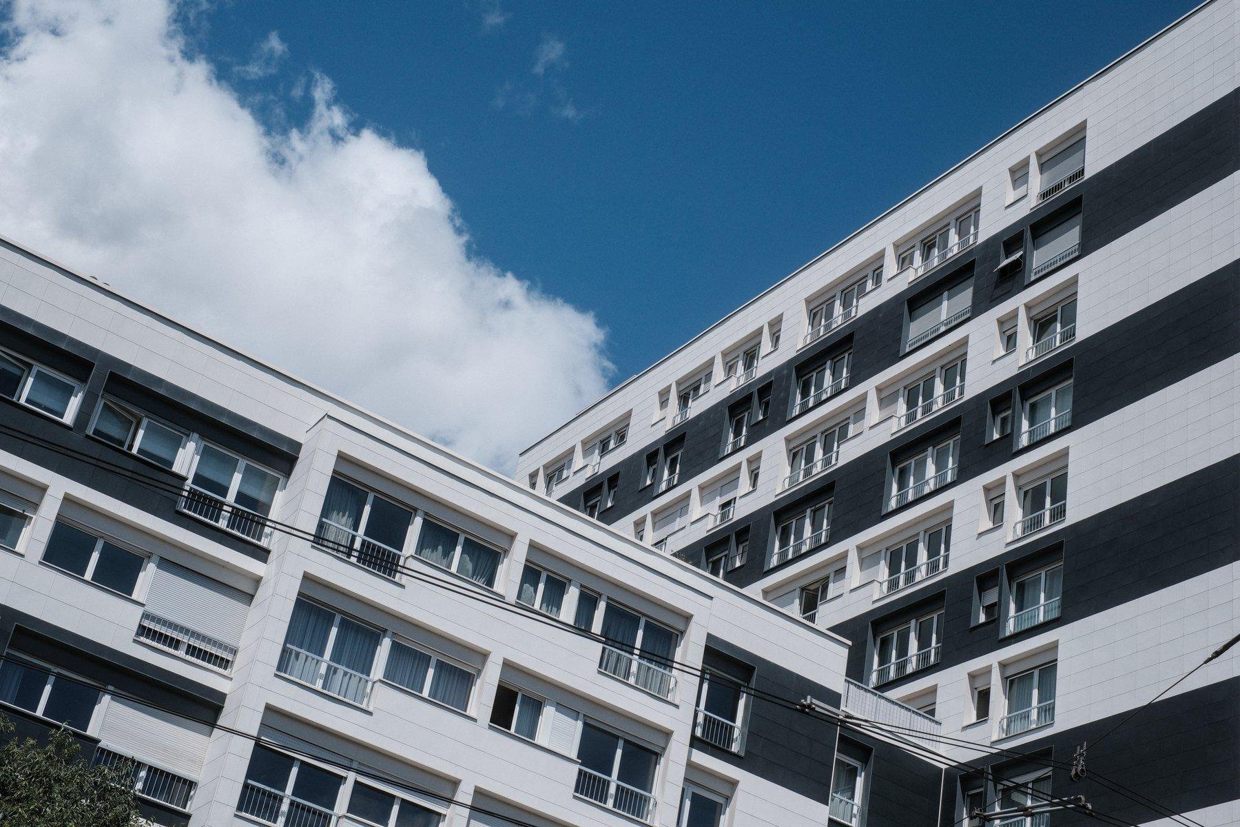 Photo horizontale. À droite, un immeuble noir et blanc, un étage sur deux. Sortant de lui perpendiculairement, un autre immeuble, moins haut de 7 étages, lui aussi noir et blanc un étage sur deux et avec une colonne blanche un peu en relief occupant sûrement 2 habitations de largeur
