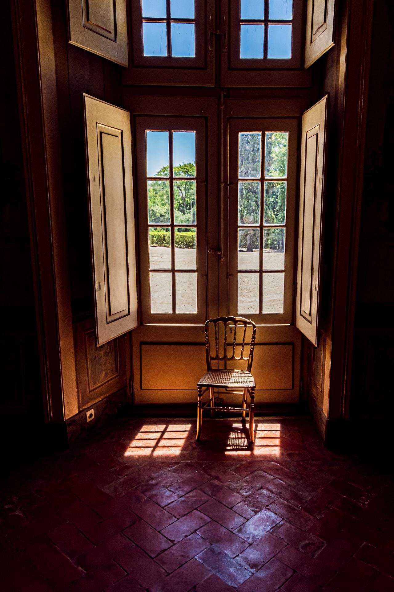 Wooden chair placed in front of tall, open double doors with large glass panes, revealing a garden view with trees. Sunlight casts shadows onto a red-tiled floor inside the dimly lit room.