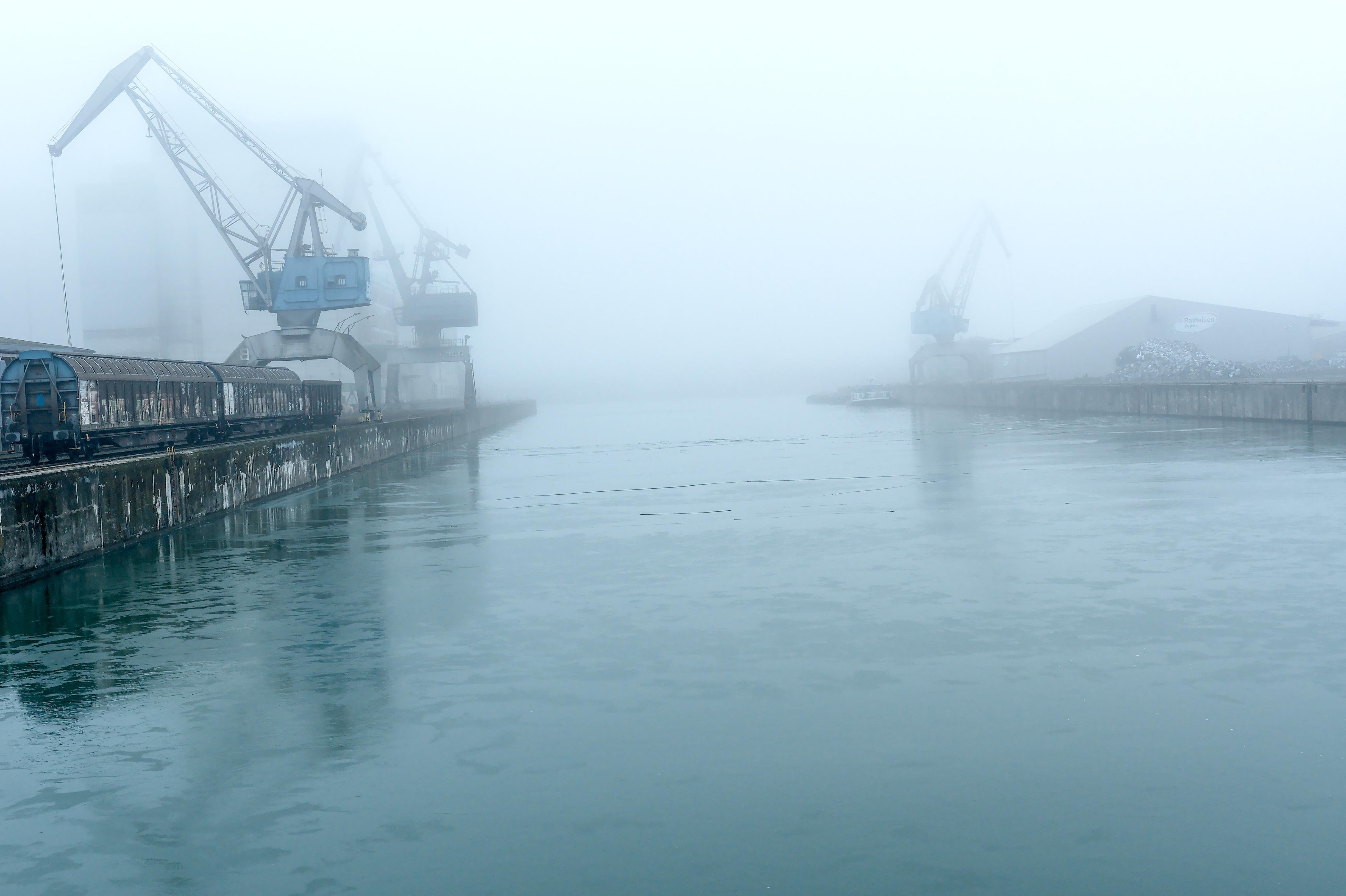 Foggy harbor canal with calm water between two concrete docks; cargo cranes and rail cars line the left dock, and a warehouse with another crane fades into the mist on the right.