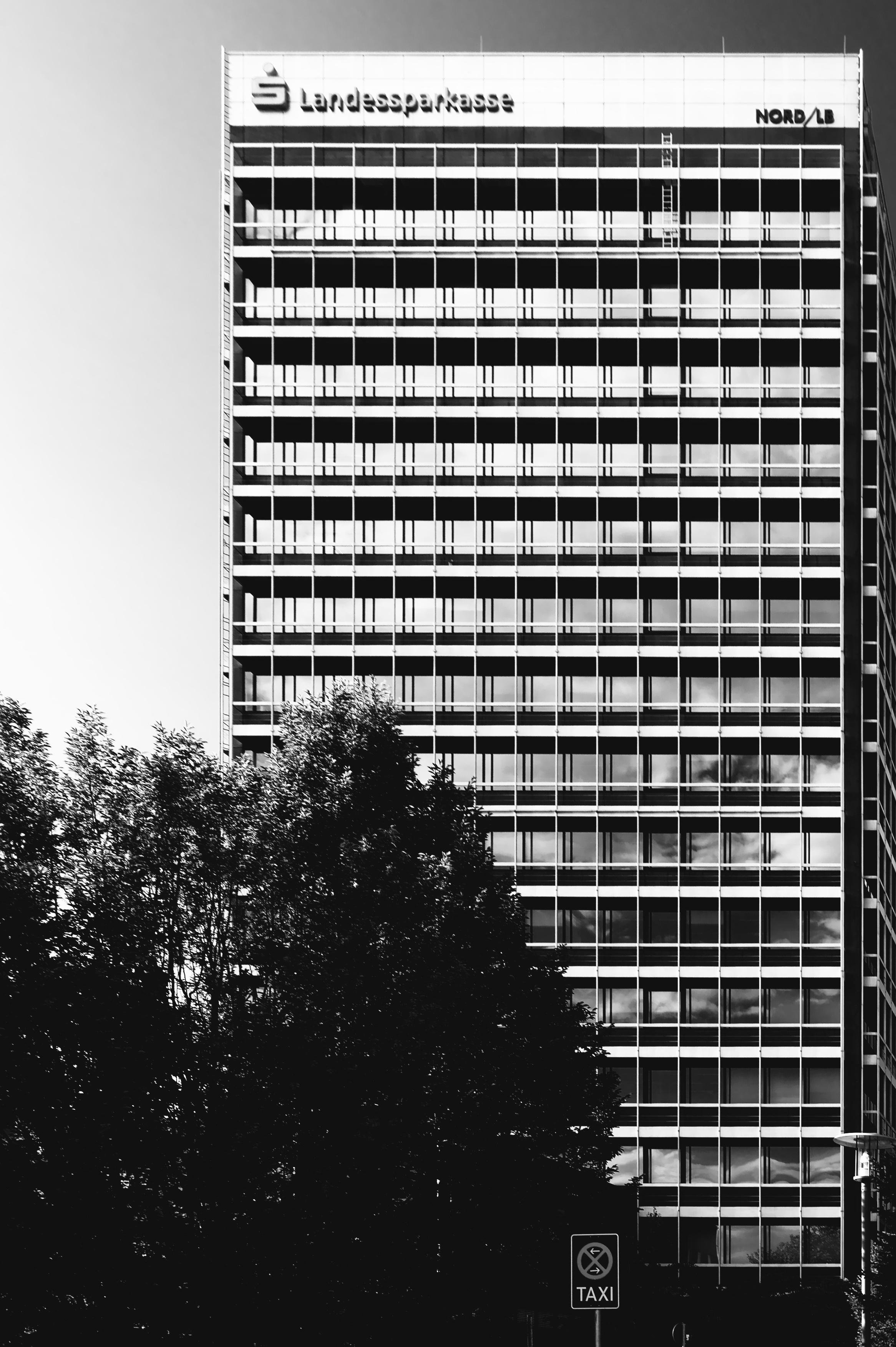 Black-and-white photo of a tall office high-rise with a grid of windows and balconies; “Landessparkasse” and “NORD/LB” signage along the top, trees in the foreground, and a small “TAXI” sign near the bottom.