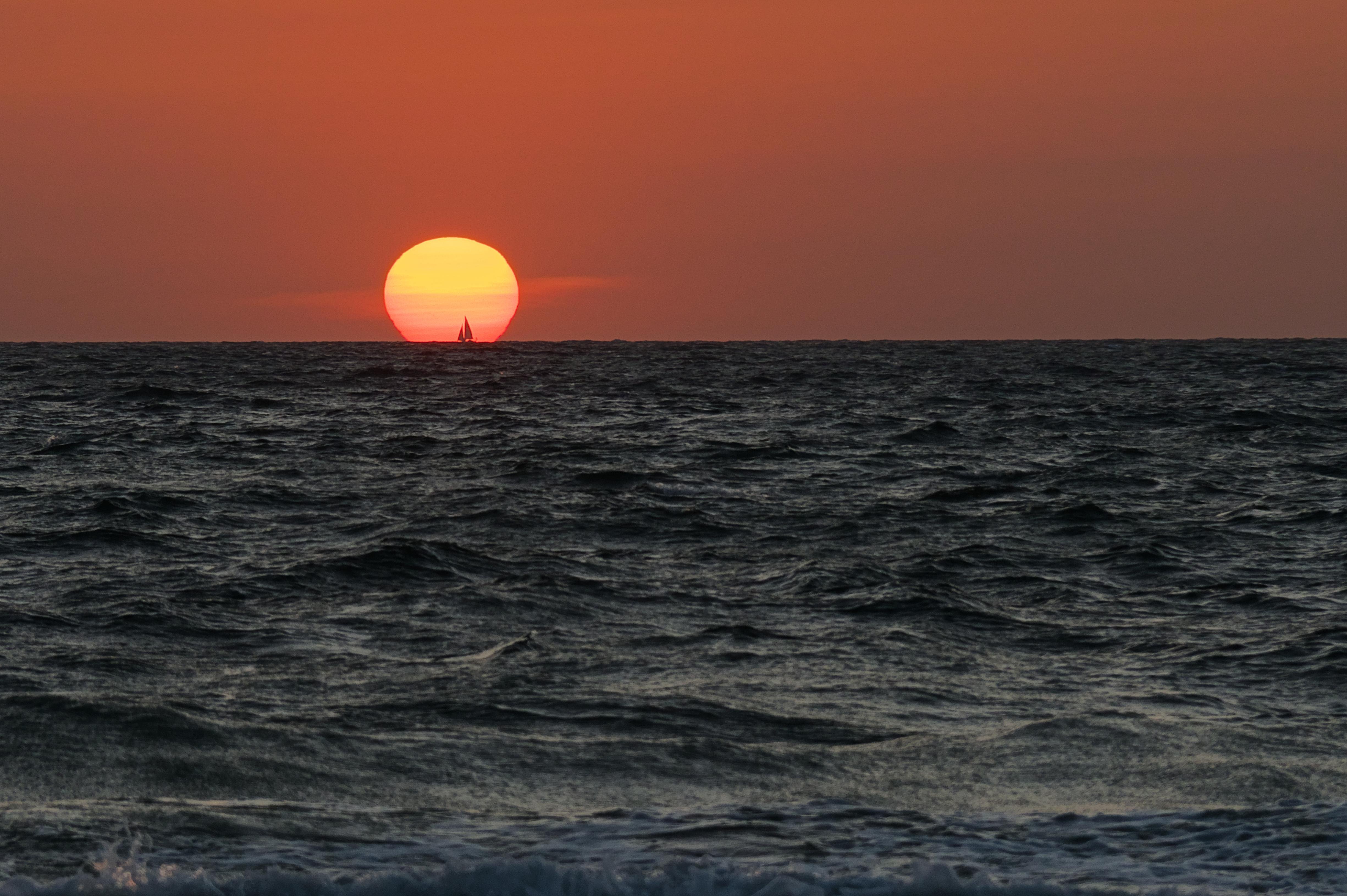 Sunset over the ocean with a large, orange sun near the horizon. A small sailboat silhouette is visible against the sun. The sky is a gradient of orange to dark, and the ocean waves are gently rolling.